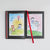 A hardcover book shown open on a light gray background, featuring two full-color childlike drawings on the pages and a red satin ribbon bookmark.